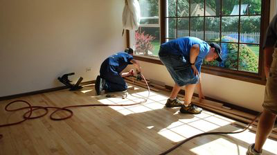 Two workers are installing wooden flooring in a sunlit room. Cables and tools are spread on the wooden floor.