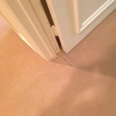 A damaged section of light brown carpet near a door frame, showing a visible tear or seam separation along the edge.