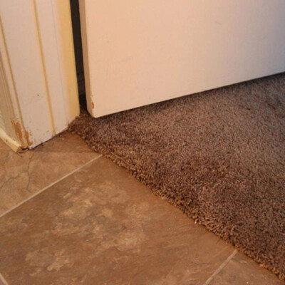 A door partially open over a floor transition with beige tile on one side and brown carpet on the other, showing wear and fraying at the carpet edge.
