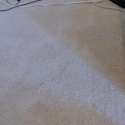 A clean beige carpet with a visible shaded area near the bottom right corner and frayed carpet sample pieces scattered on the left side. Cables are lying along the top edge of the carpet.