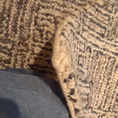 Close-up of a textured woven rug partially lifted, revealing its edge detail, with a corner of a blue fabric visible underneath.