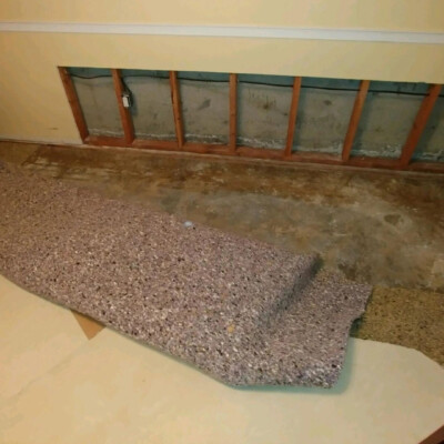 Partially removed carpet in a room reveals exposed subfloor and wall studs. A person stands to the left outside the frame.