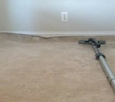 A carpet with noticeable buckling is shown with a vacuum cleaner on top, indicating an attempt to clean or flatten the surface.