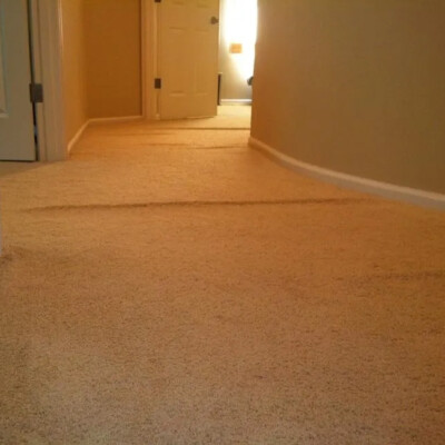 A hallway with beige carpet showing visible ripples and bumps, leading to a closed door. The walls are painted light brown.