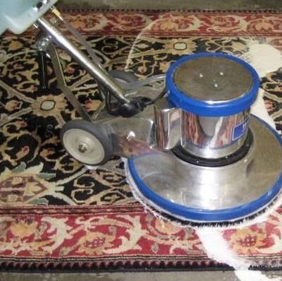 A floor-cleaning machine with a blue and silver base is being used to clean a patterned carpet, with a visible section of white cleaning solution on the carpet.