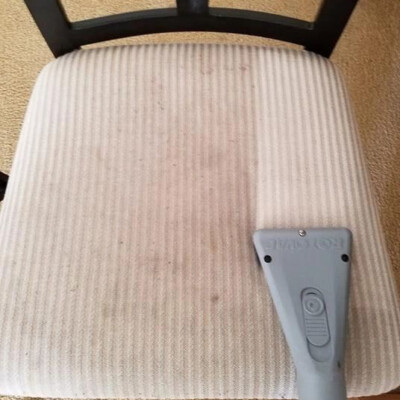 A vacuum upholstery tool is being used to clean a fabric chair seat that has visible dirt and stains.