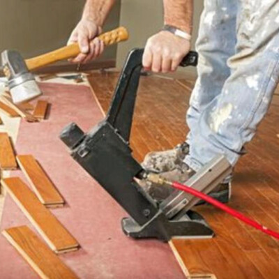 A person is installing hardwood flooring using a flooring nailer and a wooden mallet.
